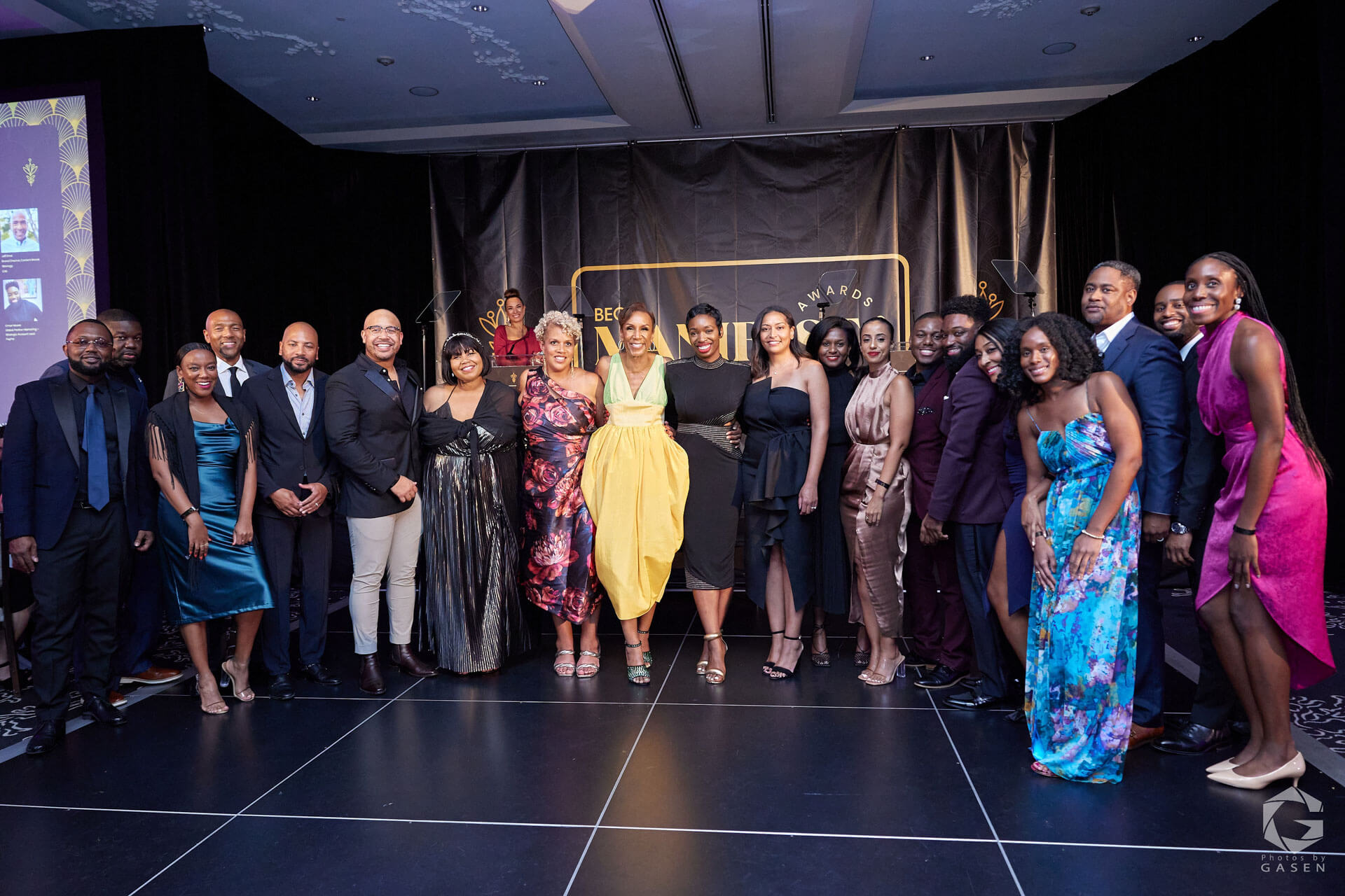 Another large group of people in formal evening wear posing on stage at an elegant event with a black-and-gold decorated backdrop.