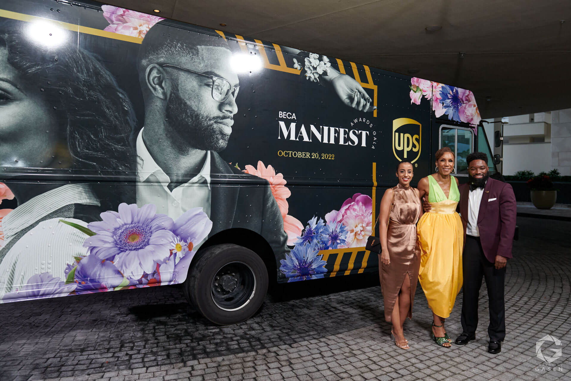 Three individuals pose in front of a floral-decorated van with "BECA Manifest Awards Gala" branding and the UPS logo.