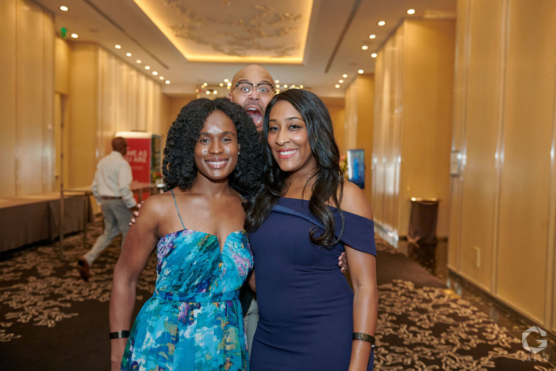 Two women dressed in elegant blue and navy dresses pose together with a photobomber playfully smiling behind them.