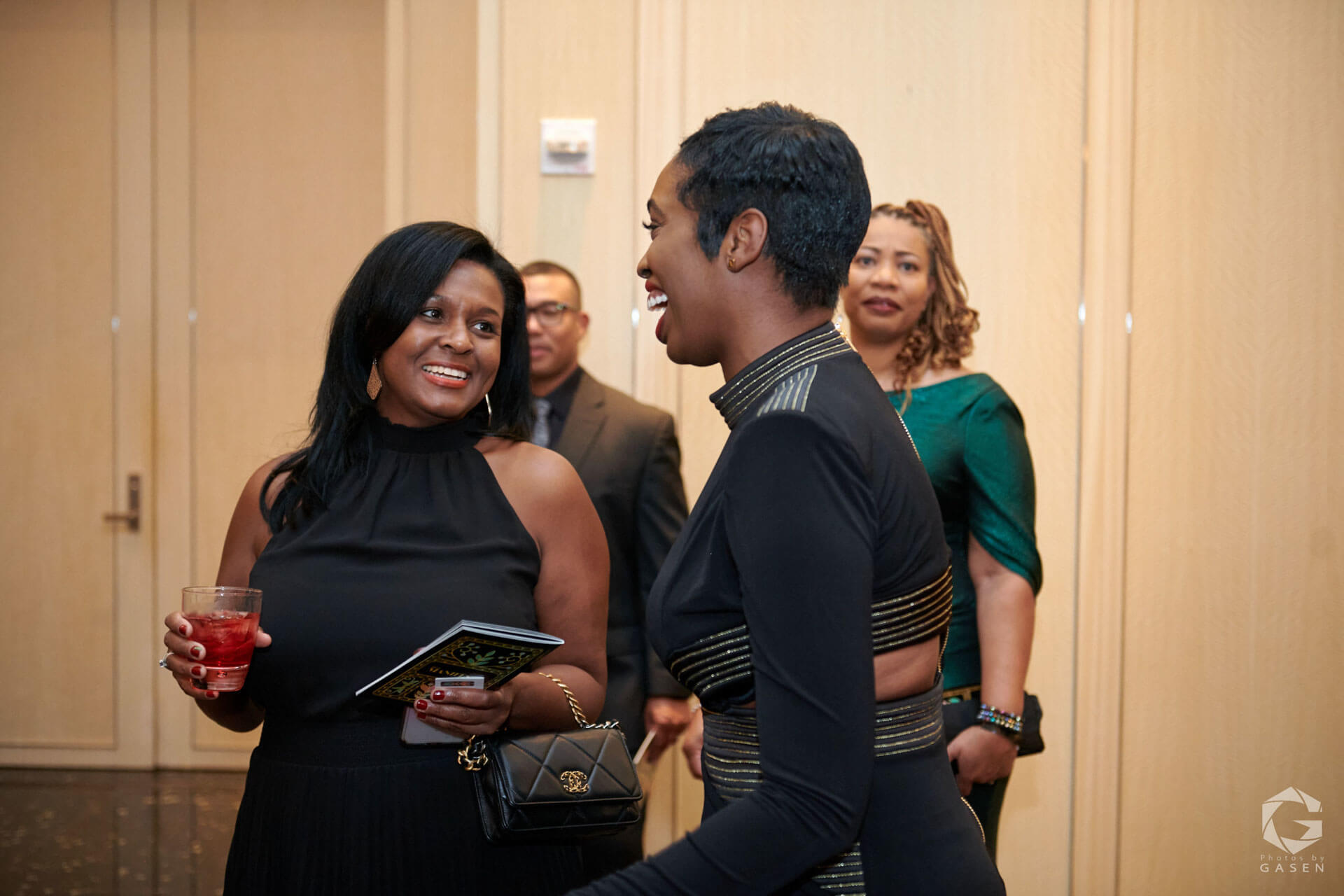 Two women in black evening attire engaged in a cheerful conversation, with other attendees and event decor in the background.