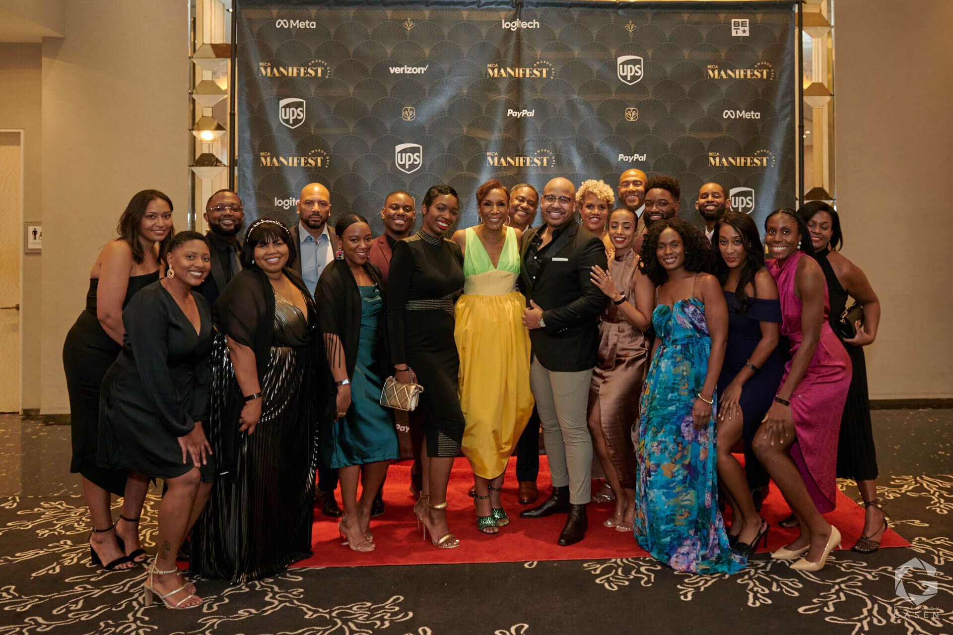 A large group of people posing for a photo on a red carpet, dressed in elegant evening wear, against a backdrop featuring event sponsors.