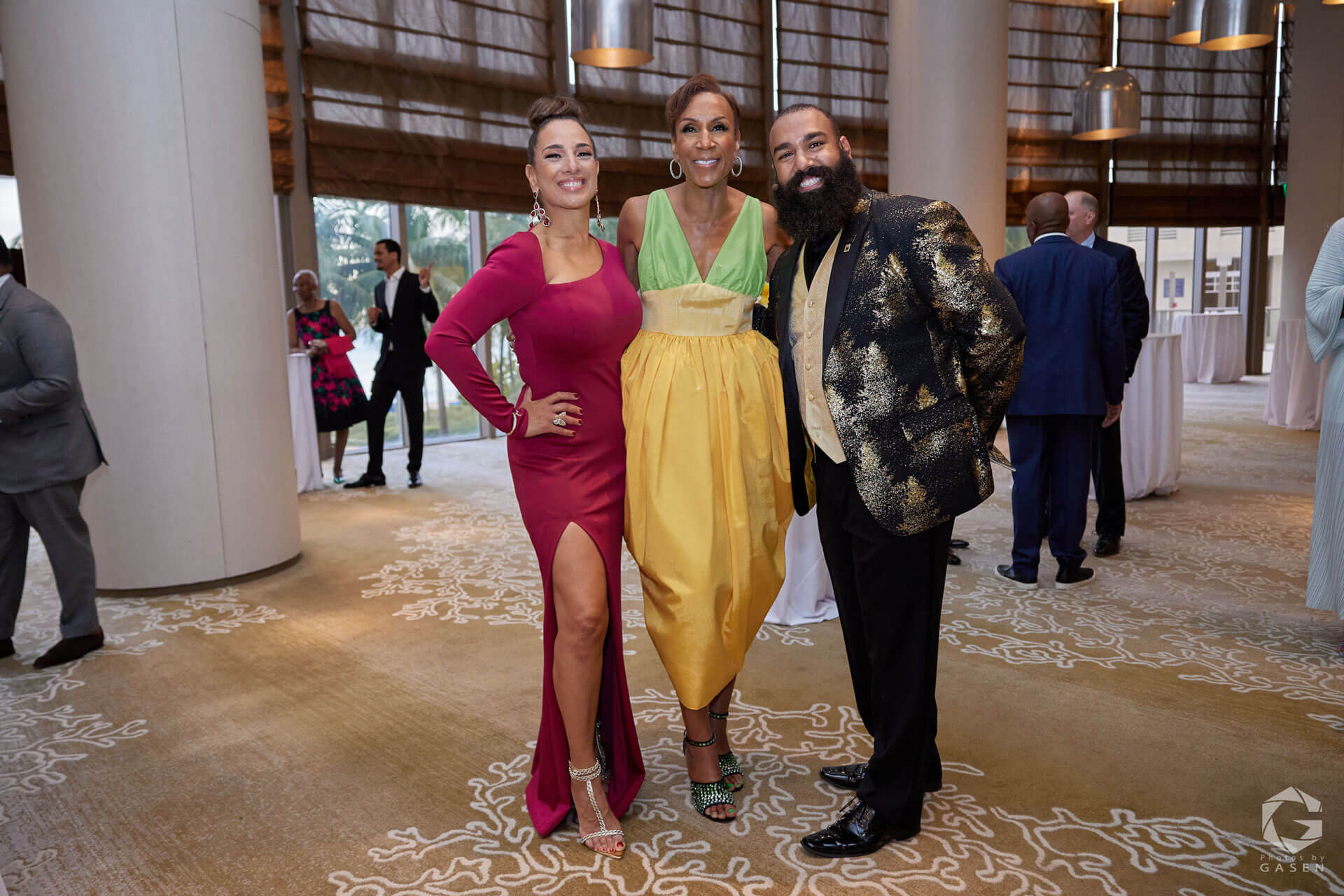 A trio posing at a formal event, including a woman in a magenta gown, another in a yellow and green dress, and a man in a gold-patterned blazer.