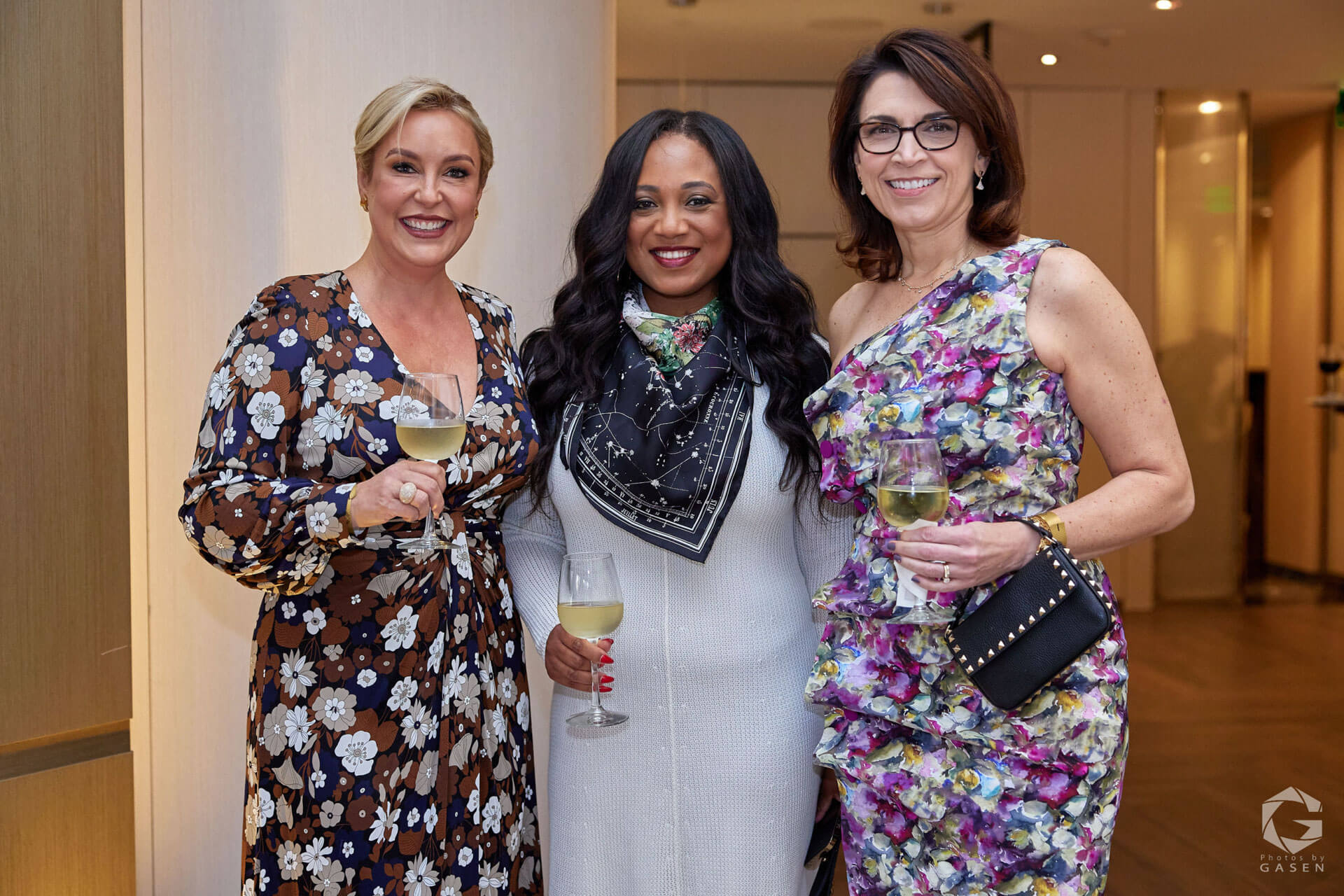Three women smiling with glasses of white wine, dressed in floral and patterned outfits, at a networking event.