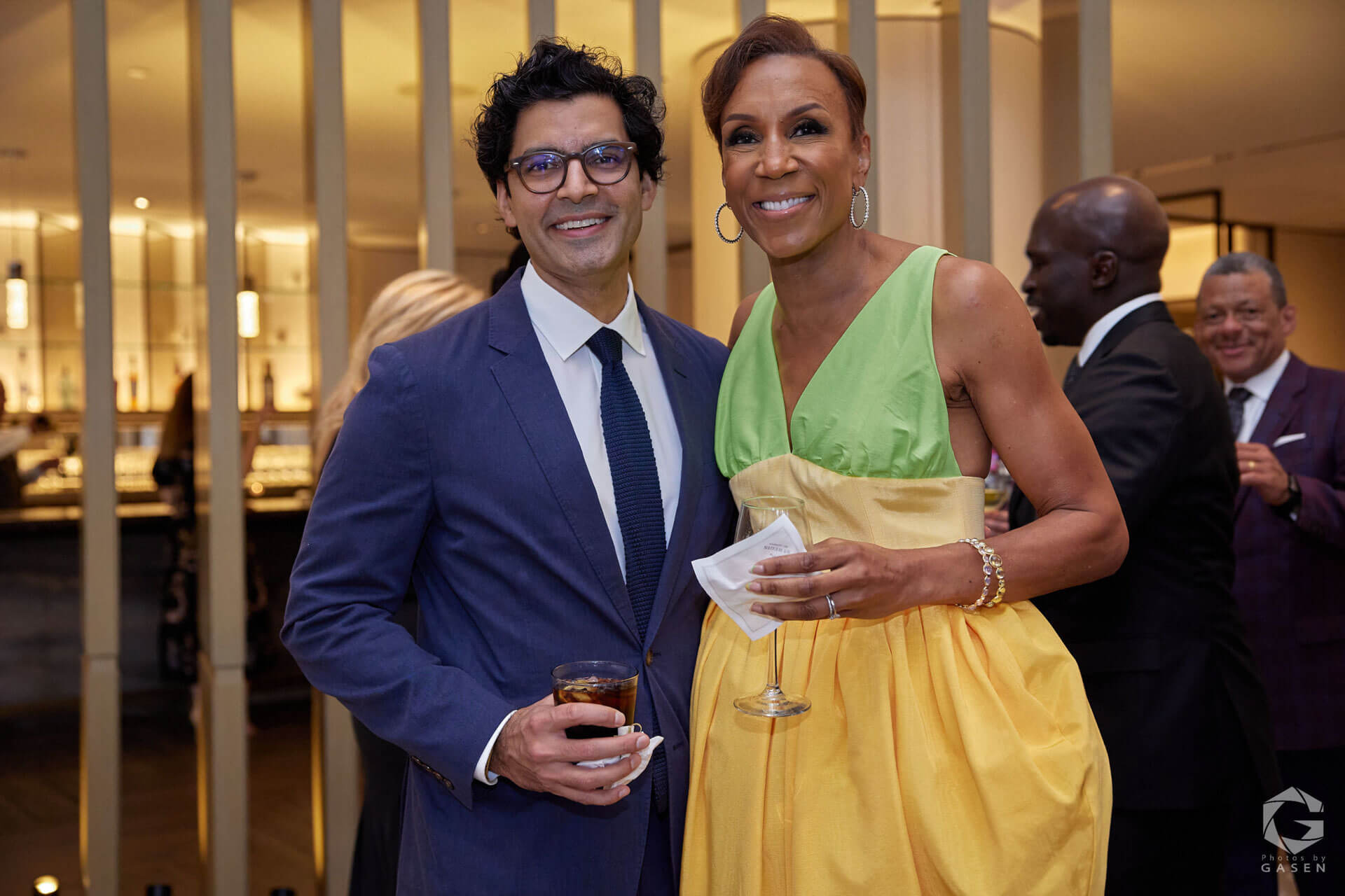 A man in a blue suit and a woman in a yellow and green dress smiling for a photo, holding drinks, in a stylish event space.