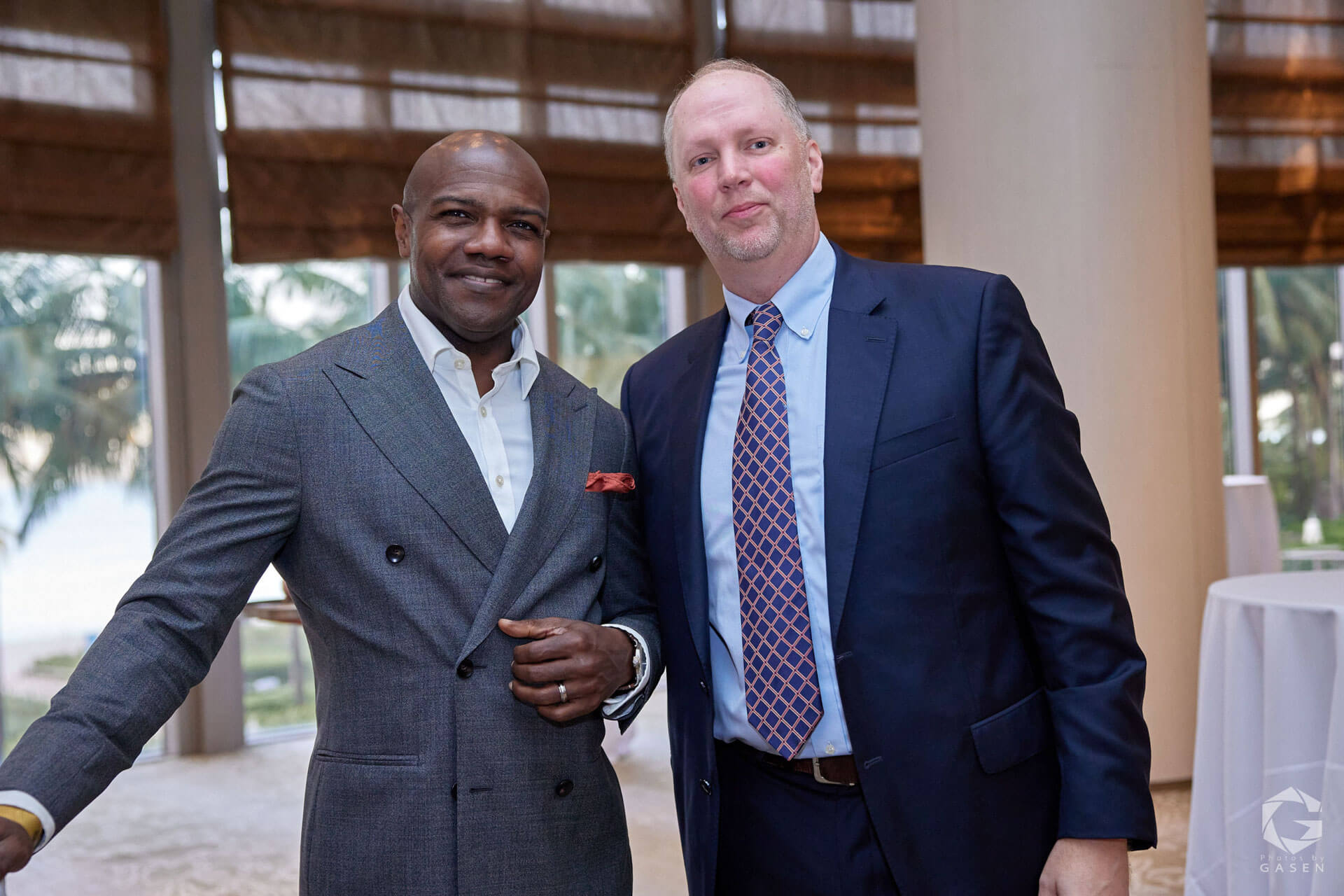 Two men in formal attire, one wearing a double-breasted gray suit and the other a navy blue suit with a patterned tie, smiling at a networking event.