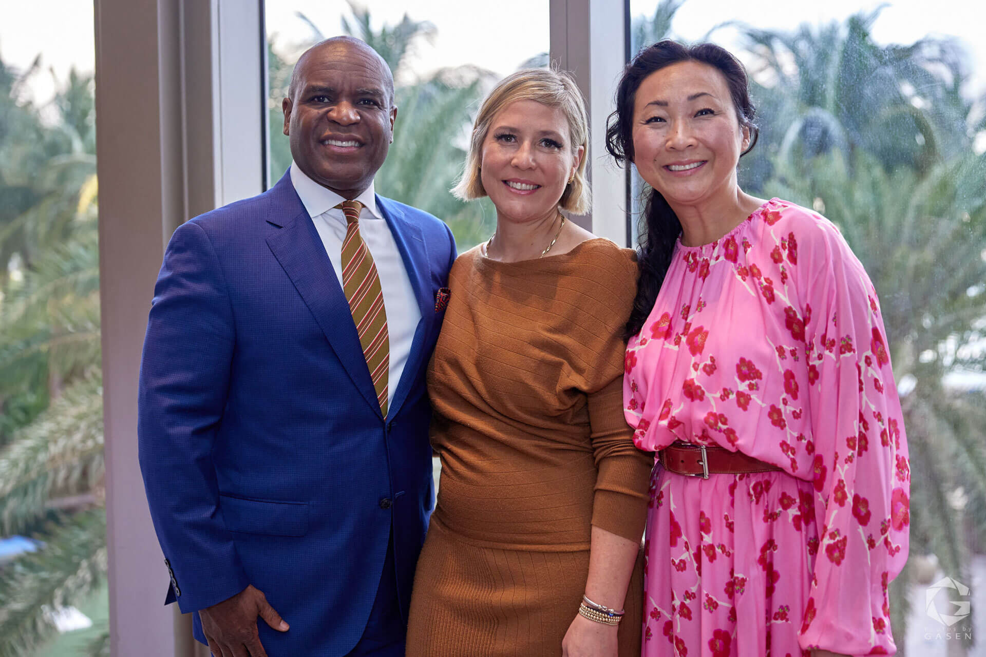 A man in a blue suit with a striped tie poses with two women, one wearing a brown dress and the other a pink floral dress, in front of a window with a tropical view.