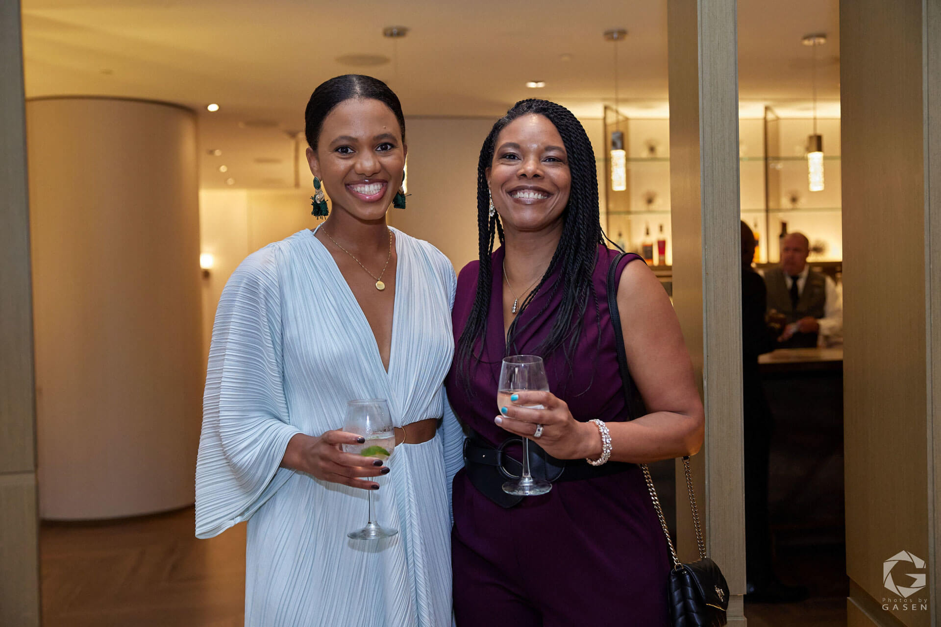 Two women smiling while holding wine glasses, one in a light blue pleated dress and the other in a maroon sleeveless jumpsuit.