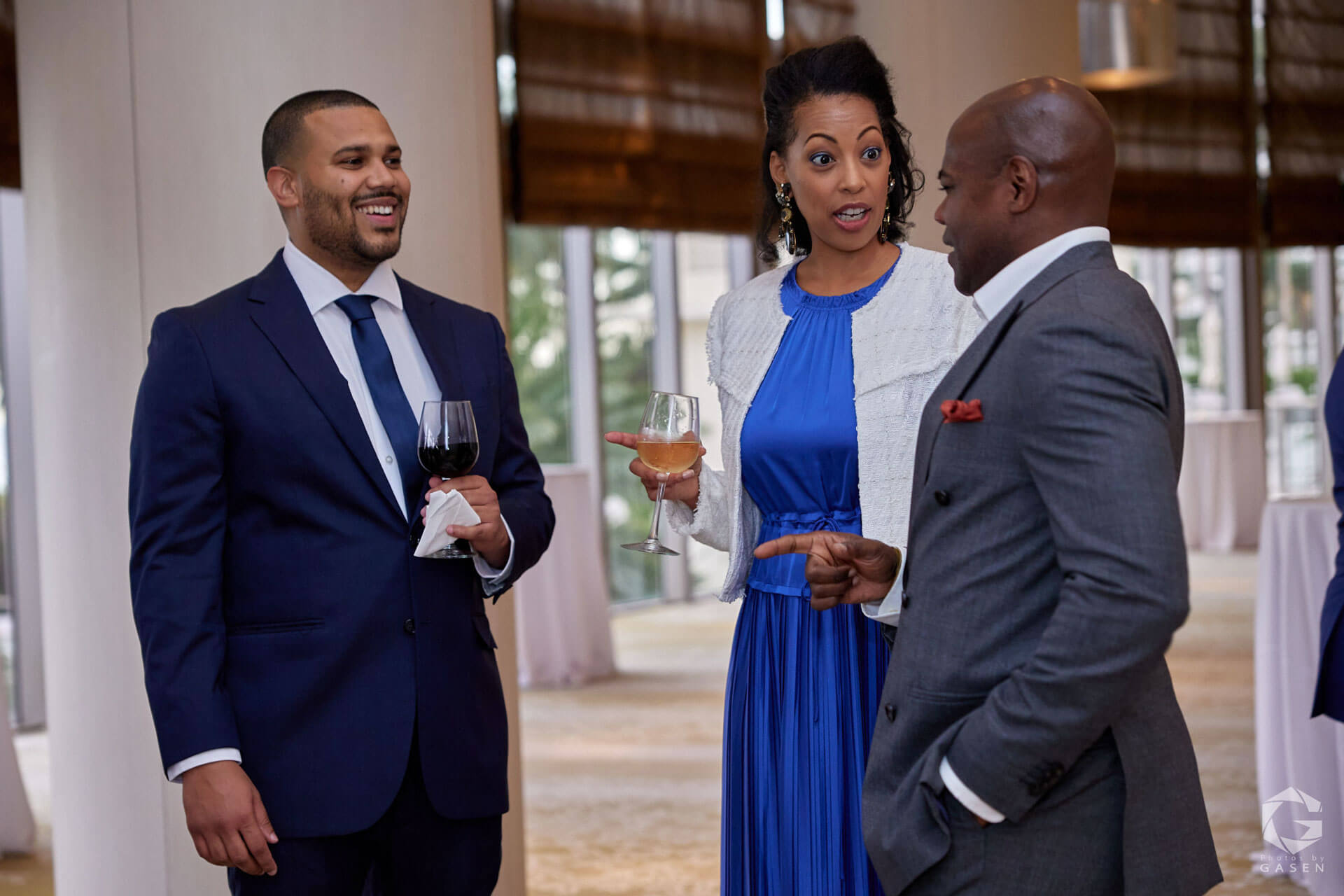 Three people engaging in conversation at an event, with a man in a suit and two women, one wearing a white jacket and a blue dress.