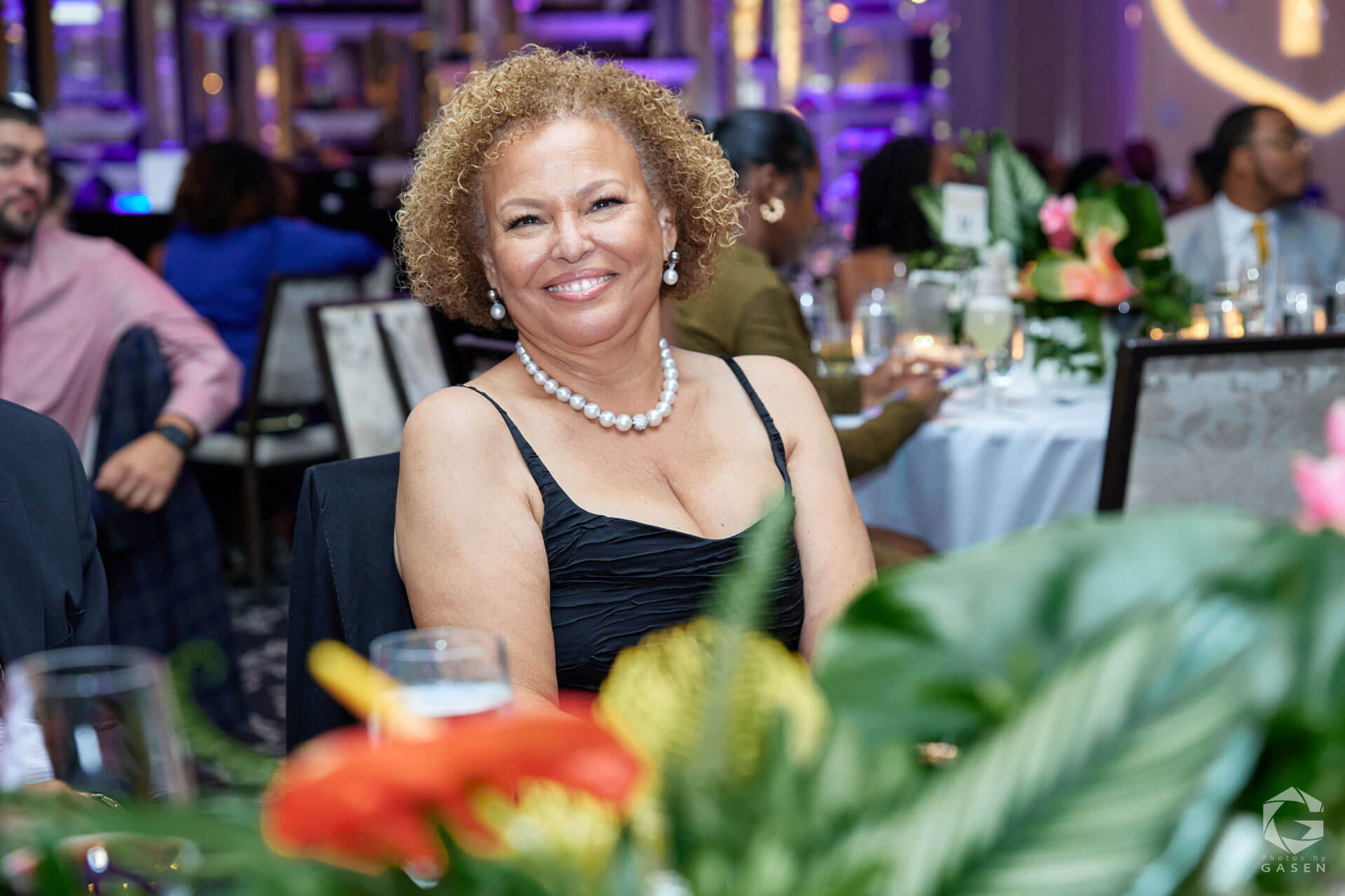 A woman with pearl jewelry and a black dress smiling while seated at a decorated dinner table.