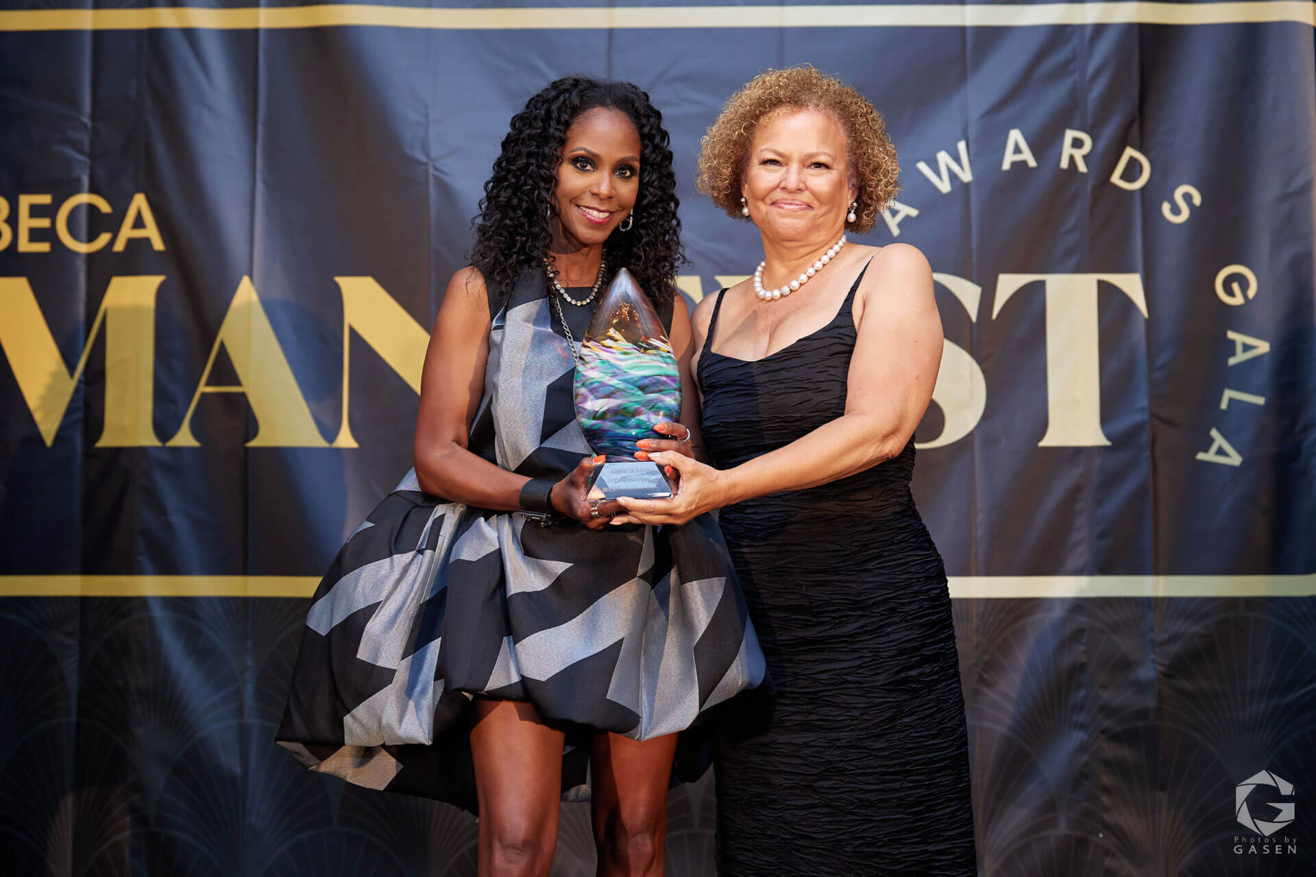 Two women stand on a stage holding an award together; one is wearing a patterned black and gray dress, and the other is in a black dress with pearl jewelry.