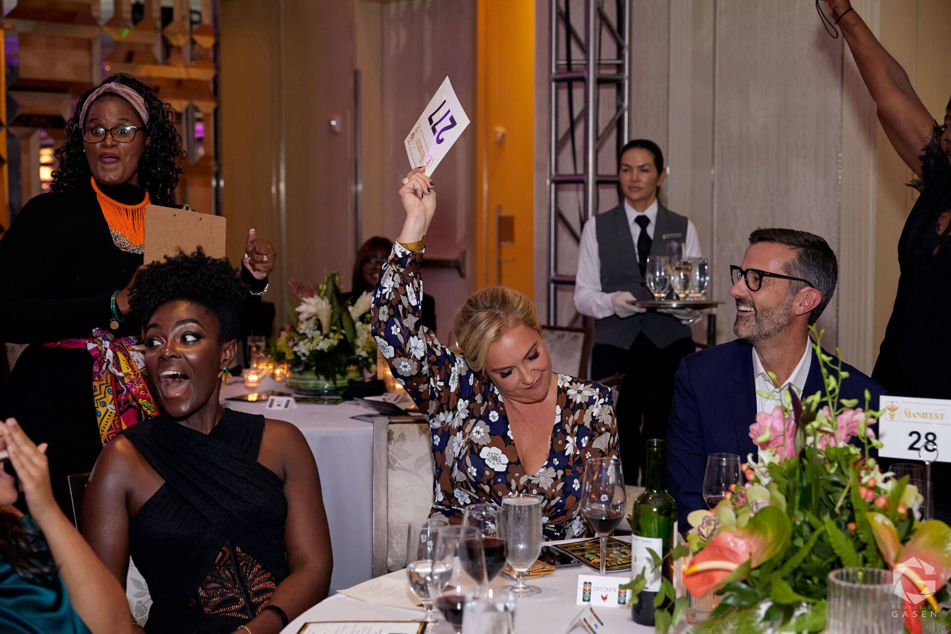 A lively auction scene with a woman holding up a bid paddle marked "211" while others around her react enthusiastically at a flower-adorned table.