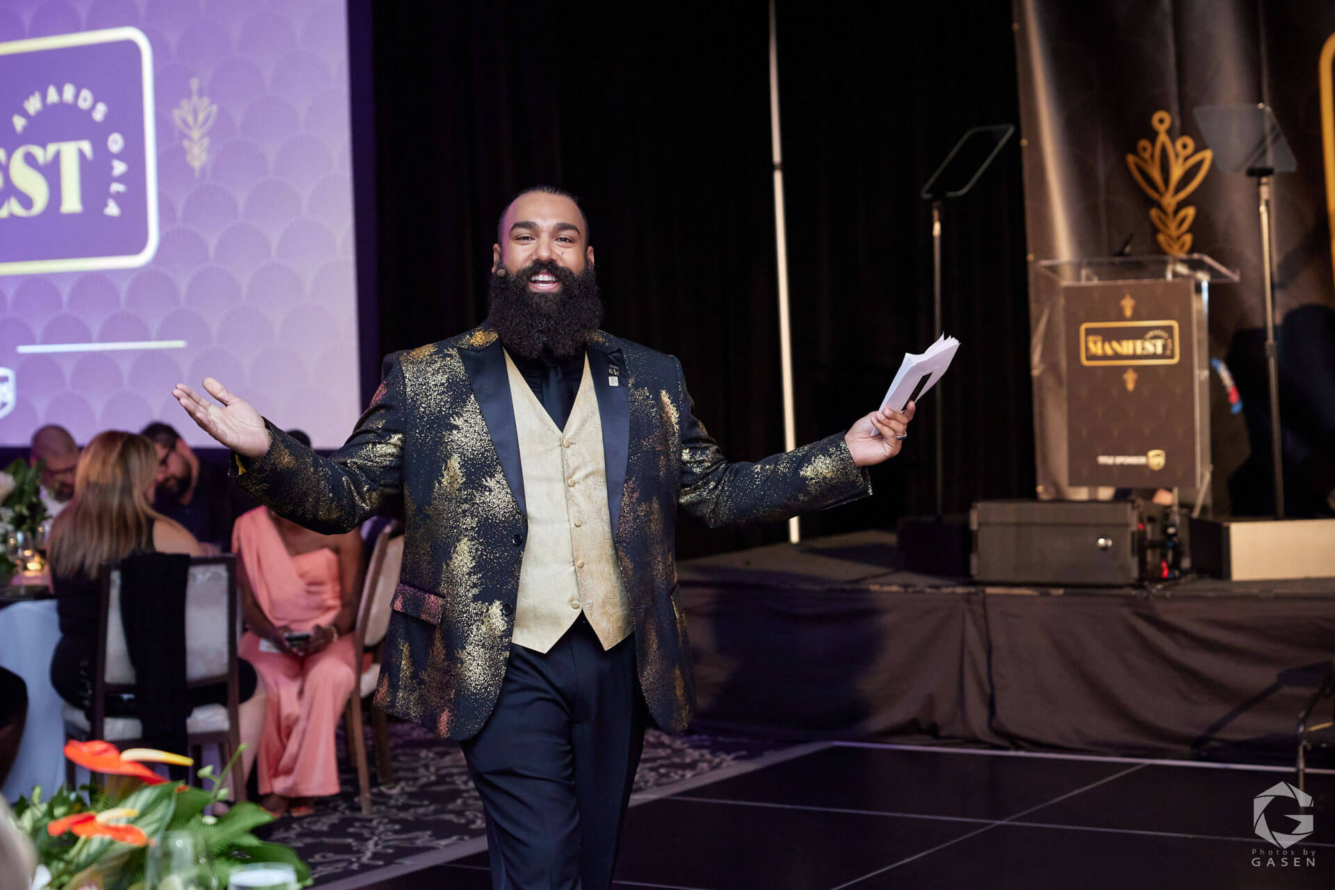 A man with a full beard and a gold-patterned jacket gestures with outstretched arms on a stage in a formal event setting.