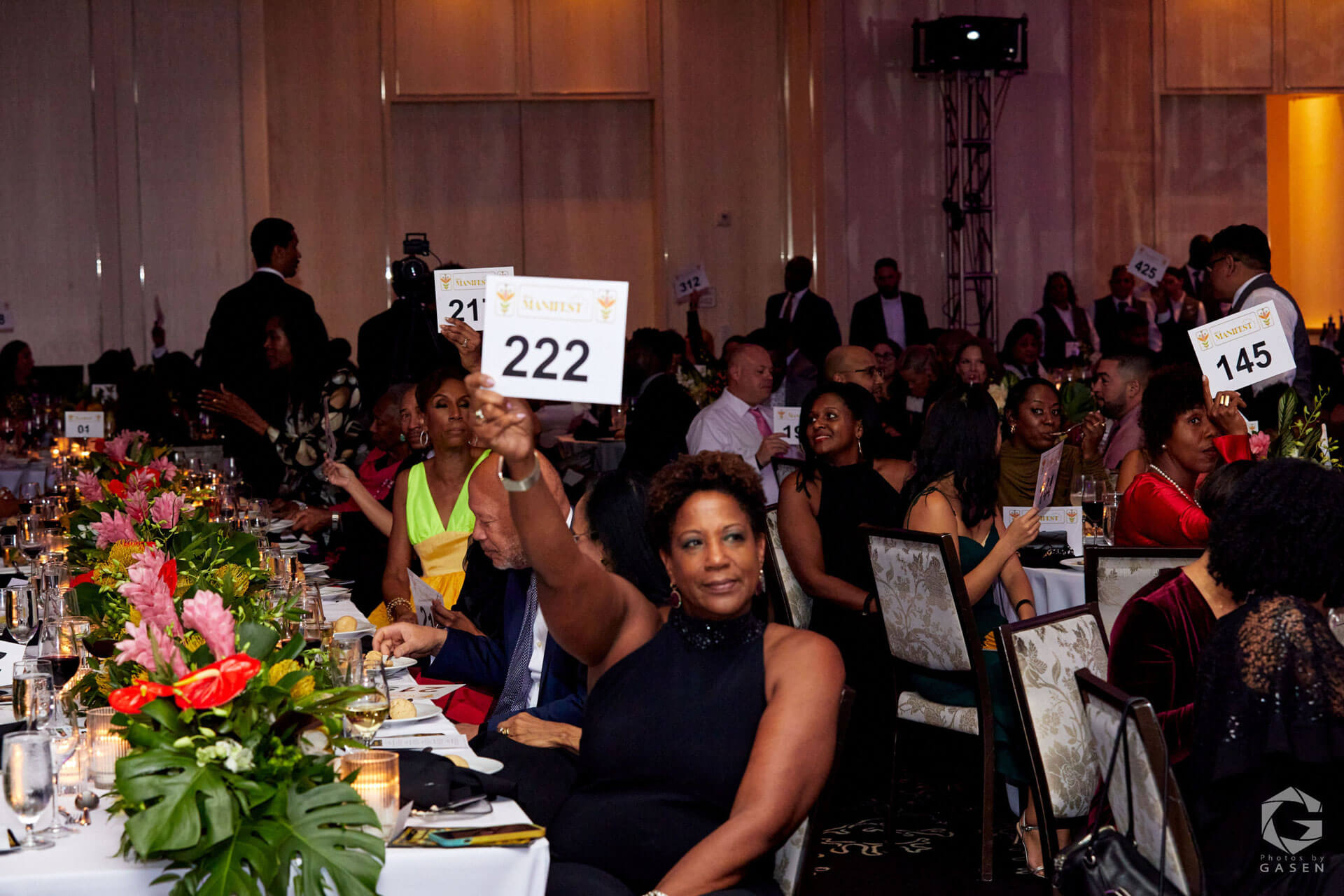 A woman in a sleeveless black dress holds up a bid paddle with "222," surrounded by other attendees at a floral-decorated dinner table.