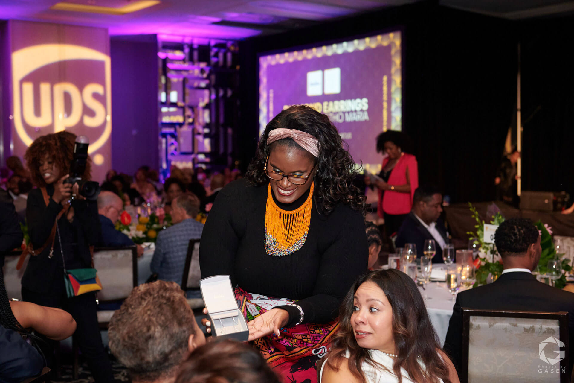A woman wearing an orange beaded necklace presents a jewelry box to guests seated at a gala table, while another woman in pink appears on stage in the background.