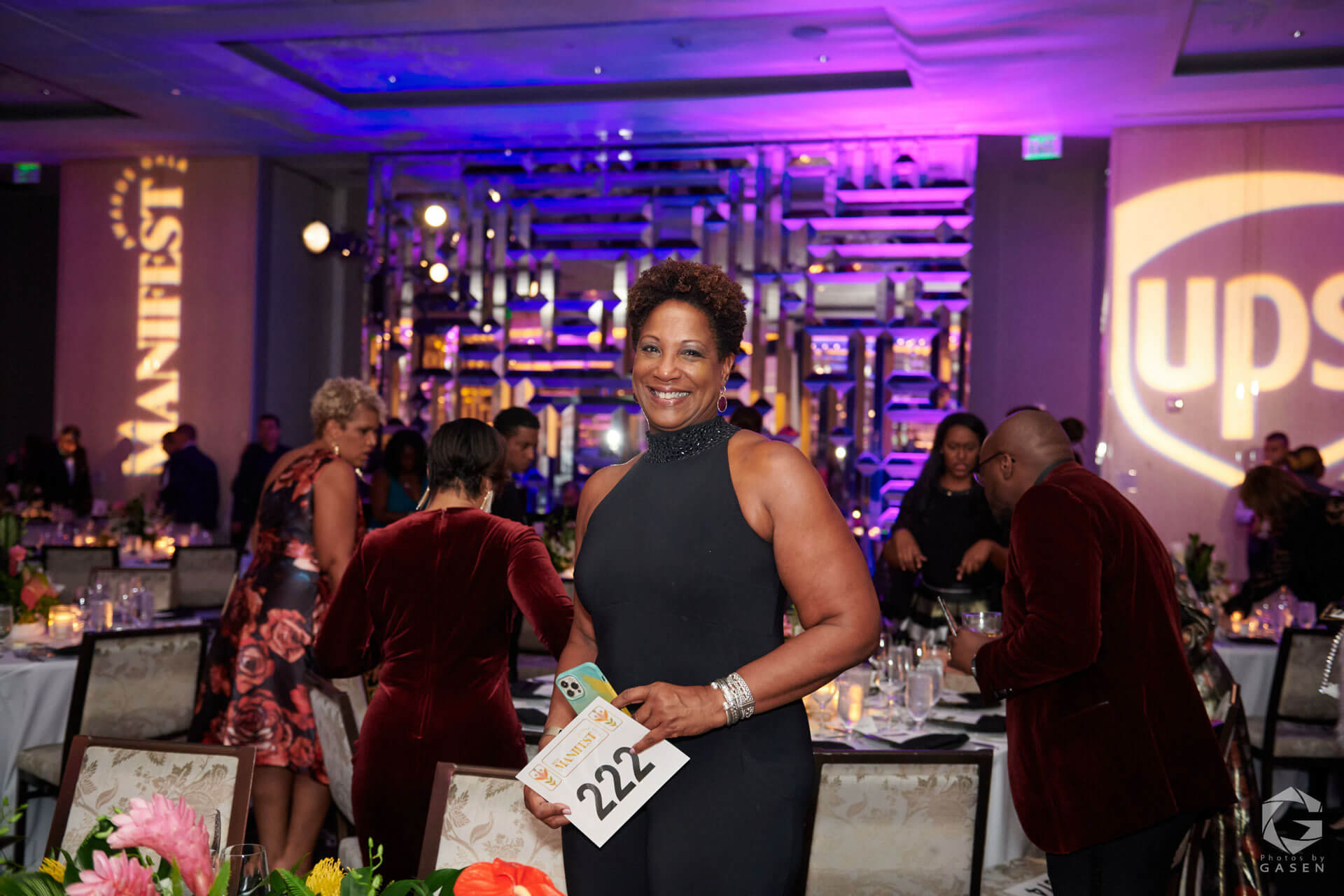 A smiling woman in a black halter gown holds a bidding paddle at a lively gala event, surrounded by decorated tables and attendees.