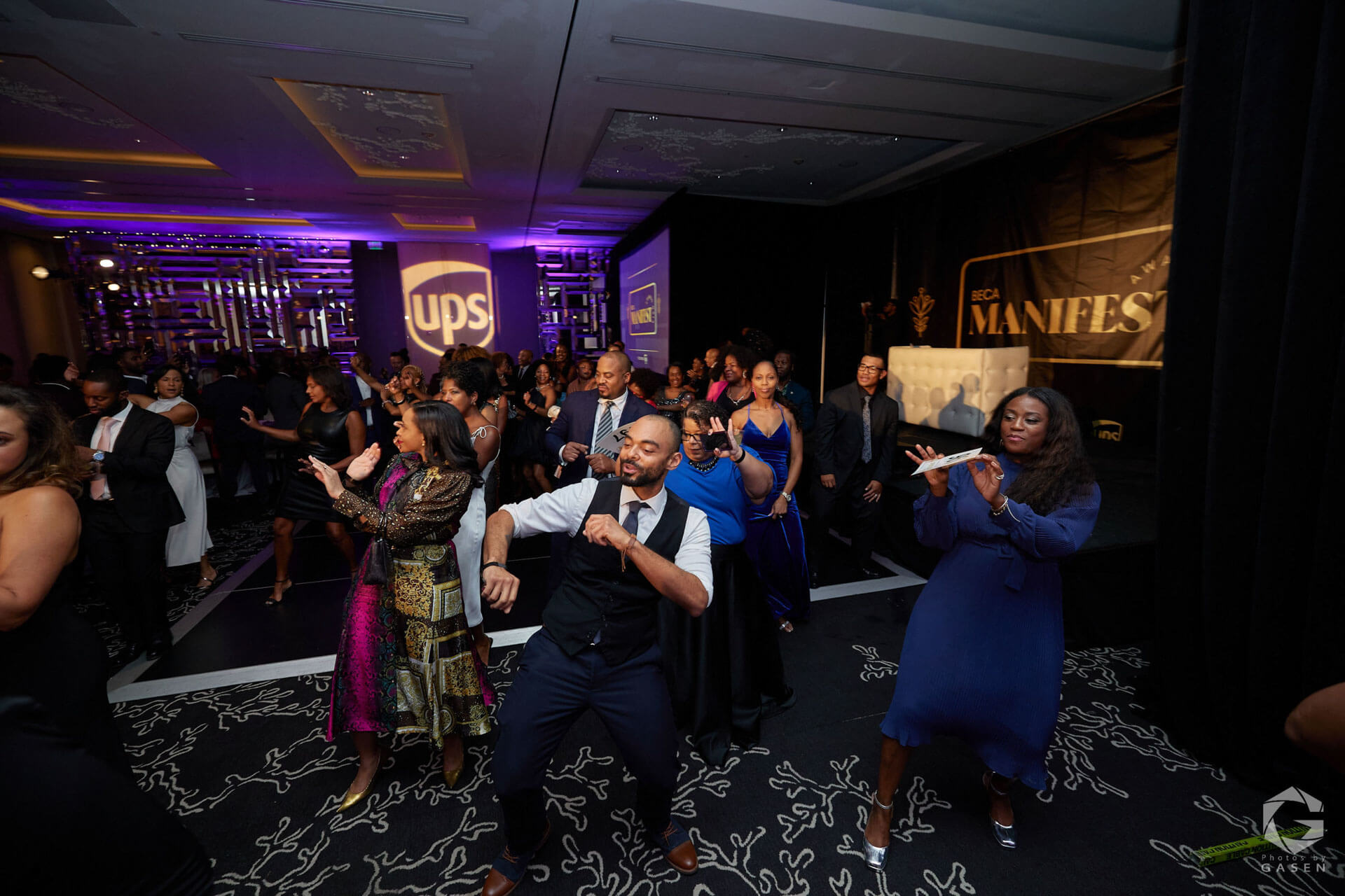 A large group of elegantly dressed attendees dance joyfully in a vibrant ballroom, with a UPS logo and "Manifest" branding visible in the background.