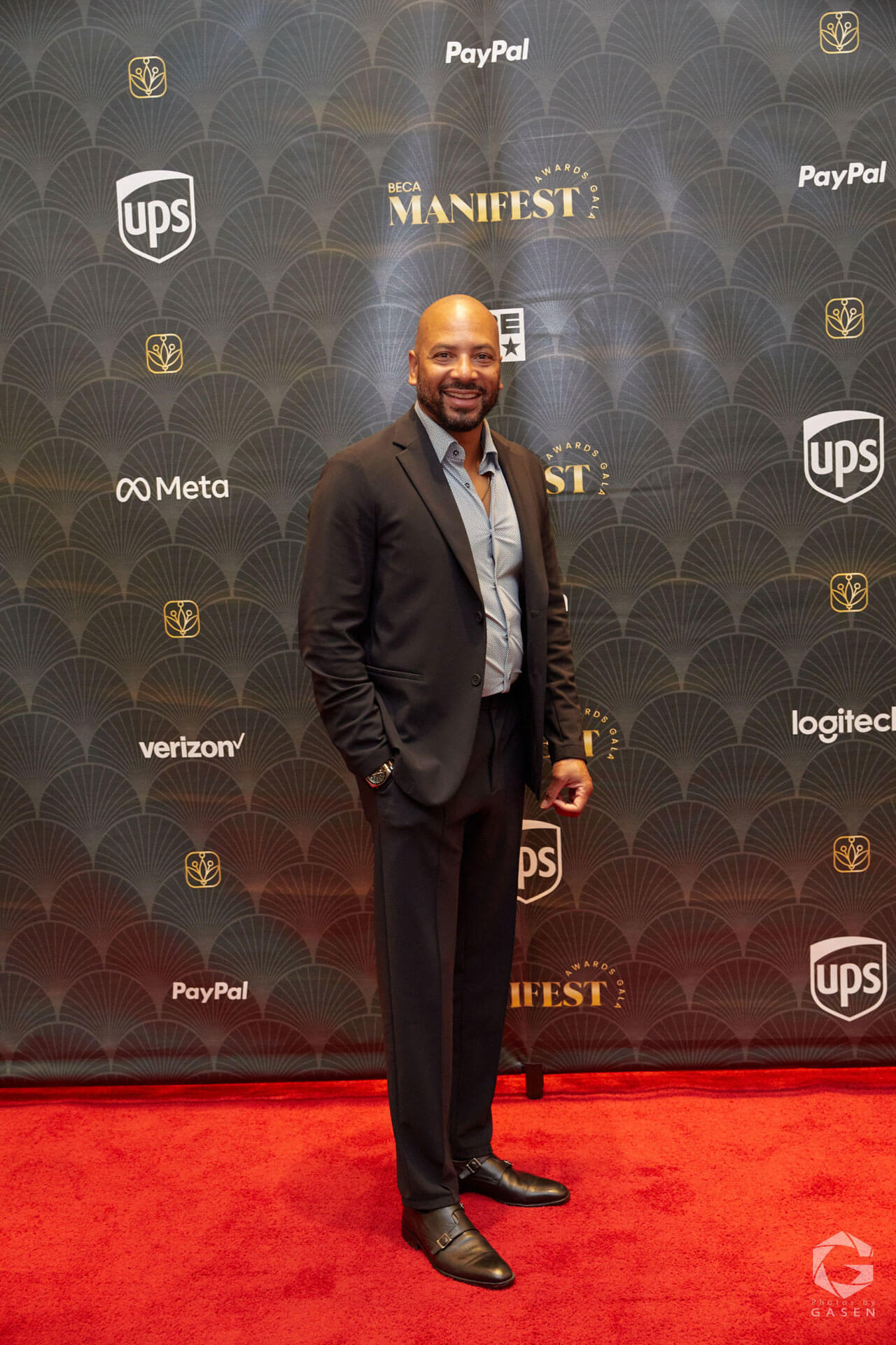 A man in a black suit and light blue shirt poses confidently on the red carpet.