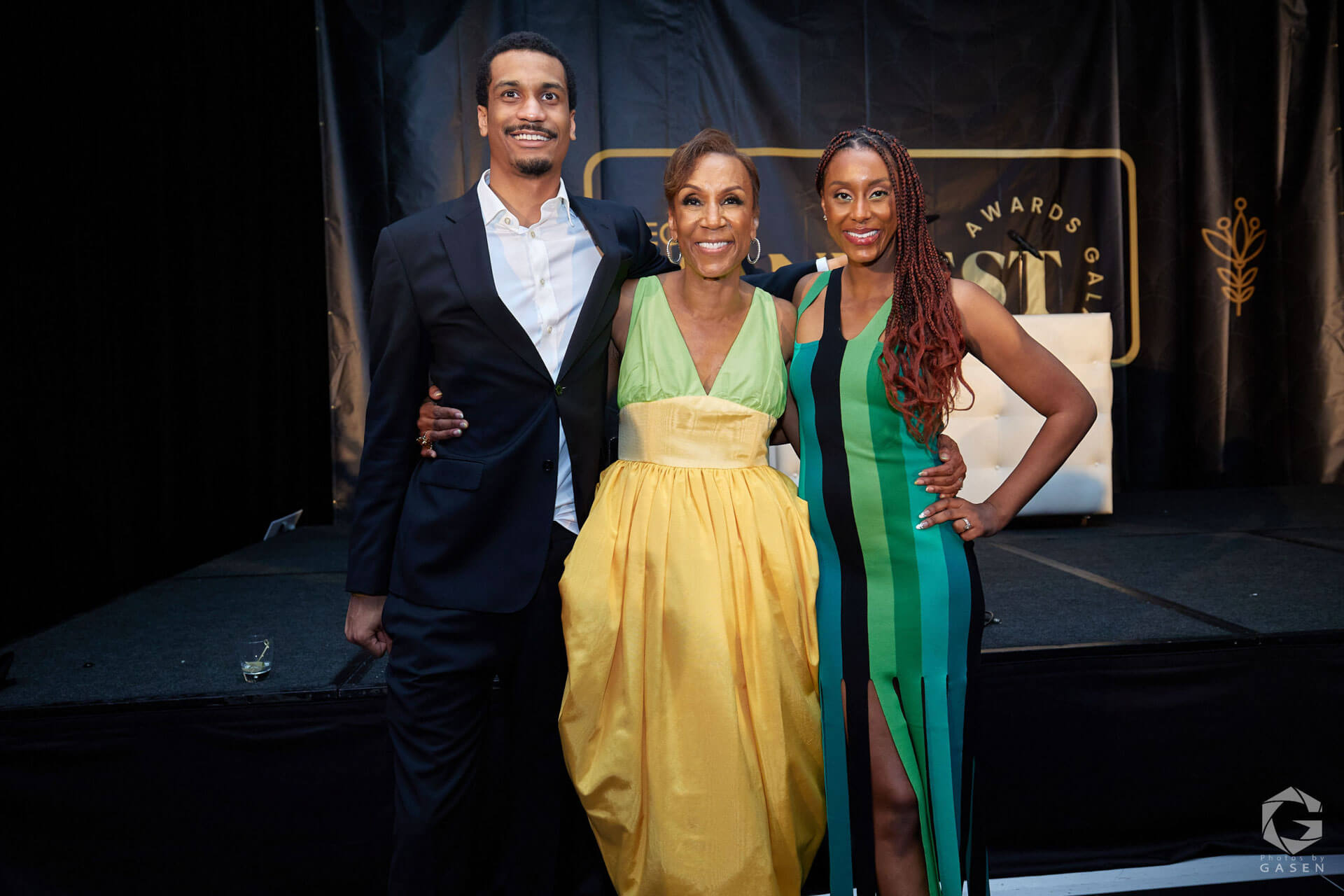 A group of three individuals posing together on a stage, with the woman in the middle wearing a vibrant yellow and green gown, flanked by a man in a suit and a woman in a green-striped dress.