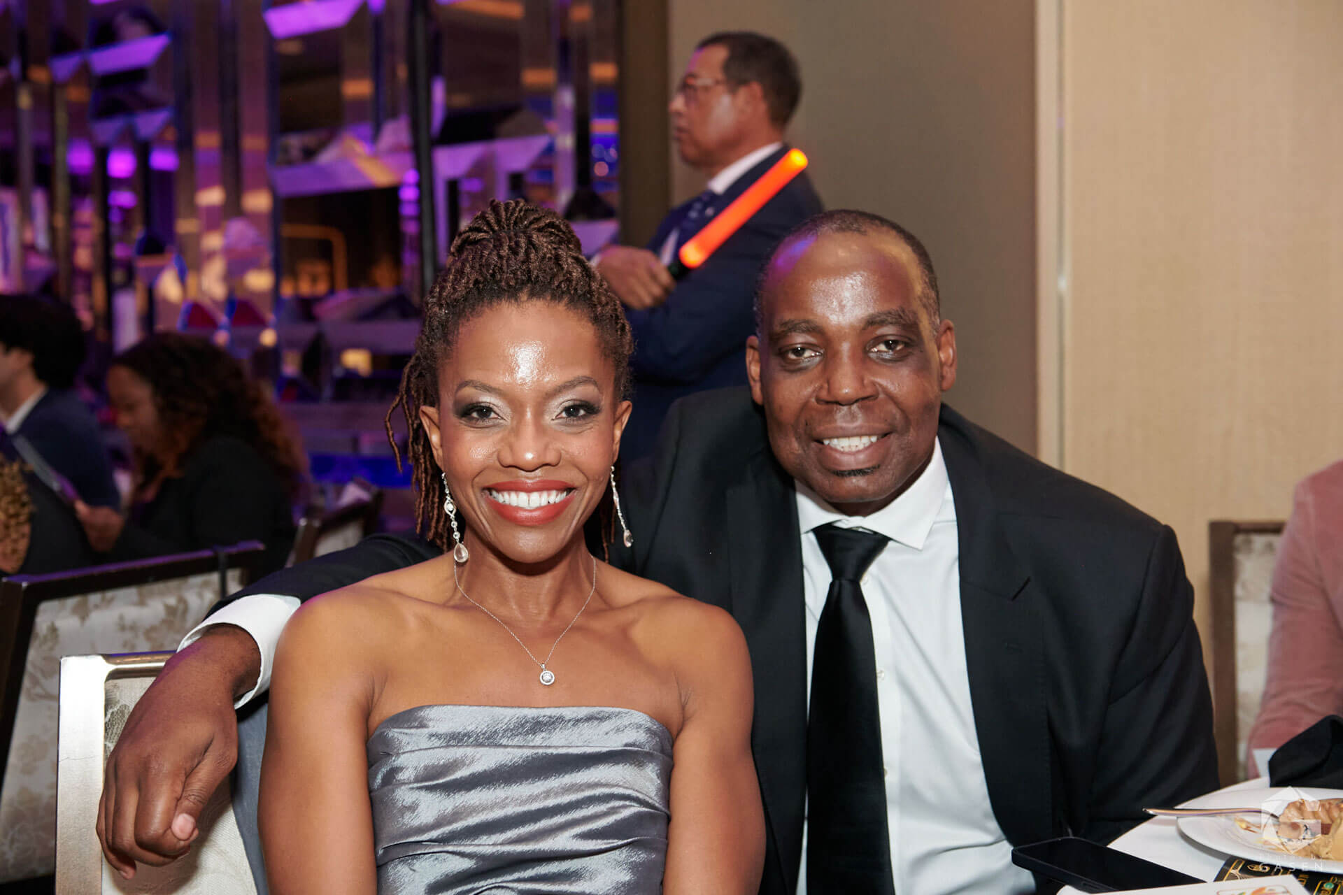 A smiling couple seated at a formal event, with the woman in a silver strapless gown and the man in a black suit and tie.