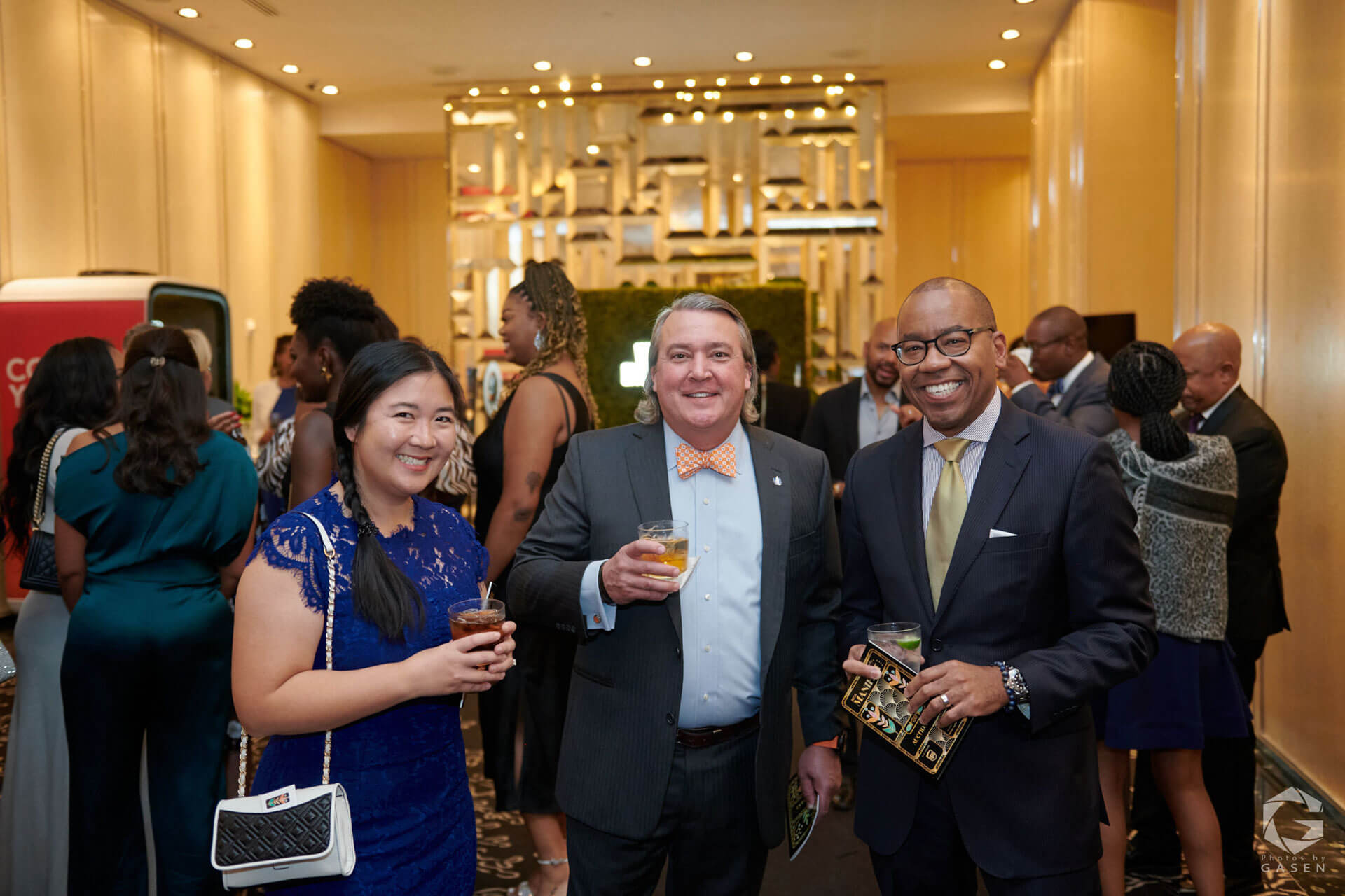 Three people, two men in suits and a woman in a blue lace dress, smiling while holding drinks at a networking event with a busy crowd in the background.
