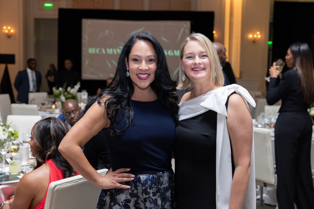 Two women pose together at a gala event, both dressed in stylish formal attire. One wears a navy blue sleeveless gown, and the other wears a black dress with a white bow detail draped over her shoulder. They are smiling with an event screen displaying "BECA" visible in the background, along with elegantly decorated tables and guests engaged in conversation.