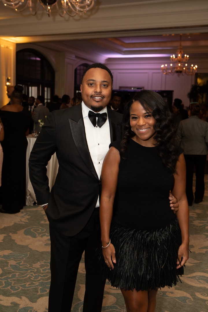 A man and a woman pose together at a formal event, both dressed elegantly. The man wears a black tuxedo with a bow tie, while the woman wears a black sleeveless dress with a feathered skirt. They both smile at the camera, with a grand ballroom and other guests mingling in the background.