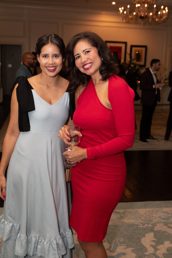Two women pose together at a formal event, smiling warmly. One wears a flowing light blue dress with black shoulder straps, while the other wears a fitted red one-sleeve dress and holds a wine glass. They stand in a sophisticated venue with chandeliers and other guests mingling in the background.