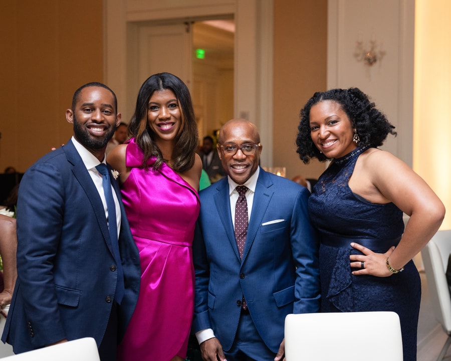 A group of four elegantly dressed individuals pose together at a formal event. Two men in navy blue suits stand alongside two women—one in a vibrant pink dress and the other in a dark blue gown. They are smiling warmly at the camera in a well-lit banquet hall with elegantly set tables in the background.