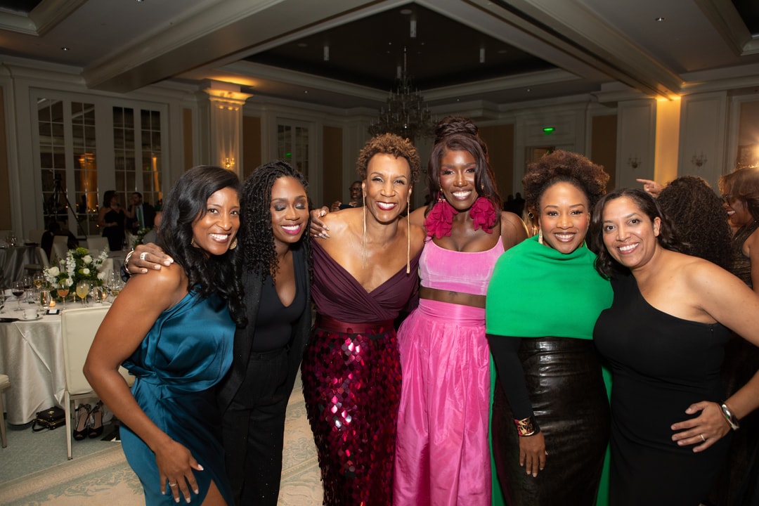 A group of six stylish women pose together at a gala event, dressed in elegant gowns and outfits in shades of blue, black, pink, and green. They are all smiling and standing closely together in a warmly lit banquet hall with chandeliers and floral arrangements visible in the background.