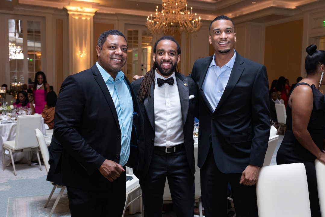 Three men stand together in a grand ballroom, dressed in formal attire. The man in the center wears a black tuxedo with a bow tie, while the other two wear dark blazers with button-down shirts. They are smiling at the camera, with elegantly set tables, guests, and chandeliers visible in the background.