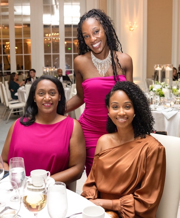 Three women are seated at a beautifully set banquet table, dressed in elegant gowns. One wears a bright pink dress, another wears a deep magenta gown, and the third wears an off-the-shoulder bronze dress. The woman in pink stands behind them, smiling brightly, while the other two are seated and looking at the camera. The background features a grand event hall with floral arrangements and chandeliers.