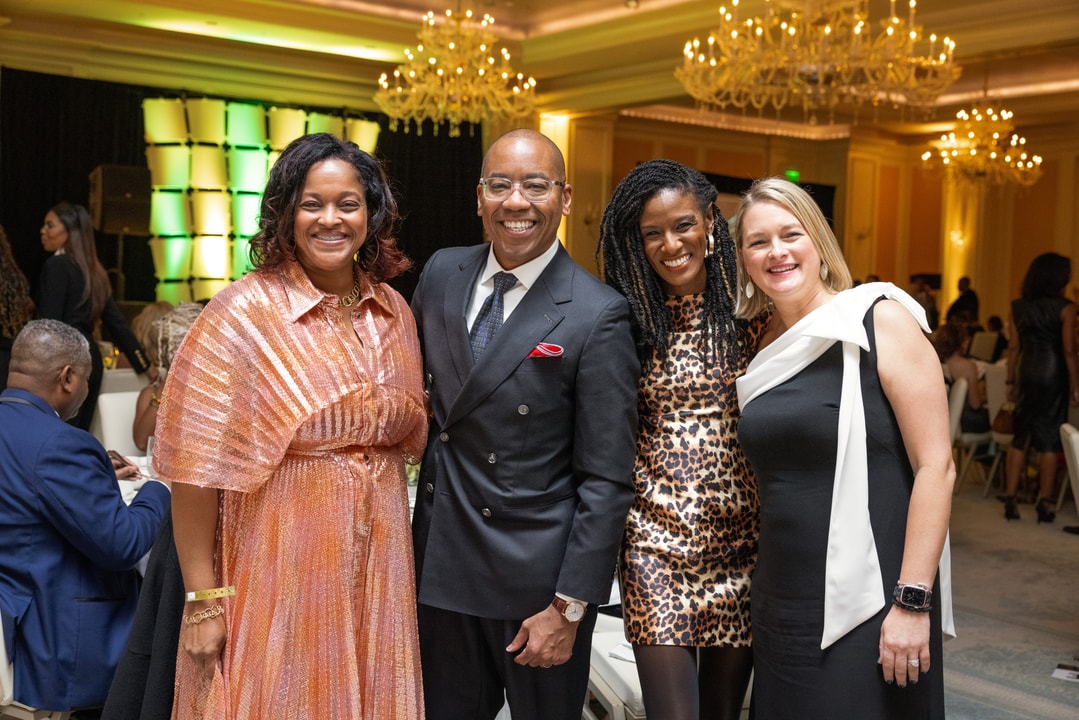 Four well-dressed individuals pose for a group photo at a formal event. The group includes a woman in a shimmering peach gown, a man in a dark double-breasted suit with a red pocket square, a woman in a leopard-print mini dress, and another in a black dress with a white shoulder bow. The elegant ballroom setting features chandeliers and an illuminated stage in the background.