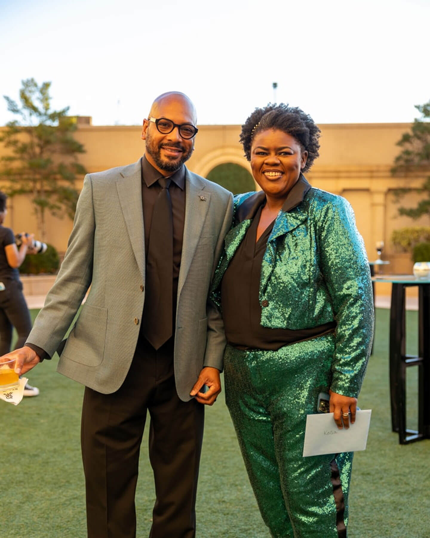 A smiling man in a tweed suit stands next to a smiling woman in a green sequined outfit at an outdoor event.