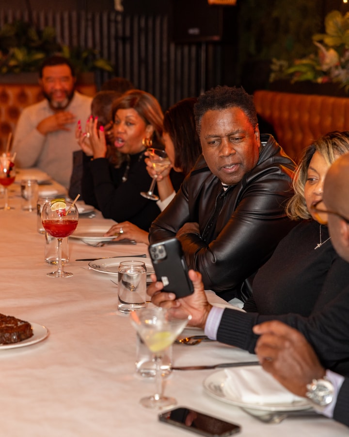 A man in a leather jacket looks at a photo shown to him on a mobile phone, while others around him talk and eat.