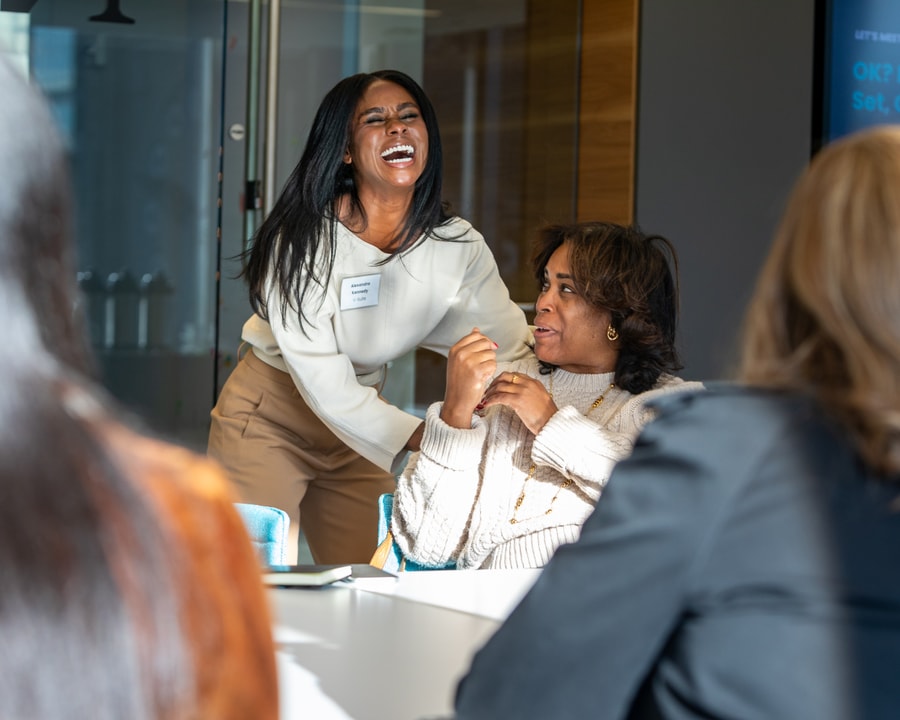 A laughing woman who is standing puts her arms around a seated woman.