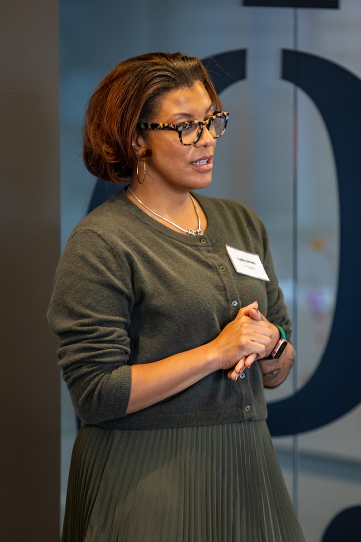 A woman in a green sweater and green skirt wearing glasses stands and presents to V-Suite Participants.
