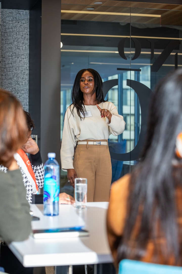 A standing woman in a white sweater and tan pants points while talking to seated colleagues.