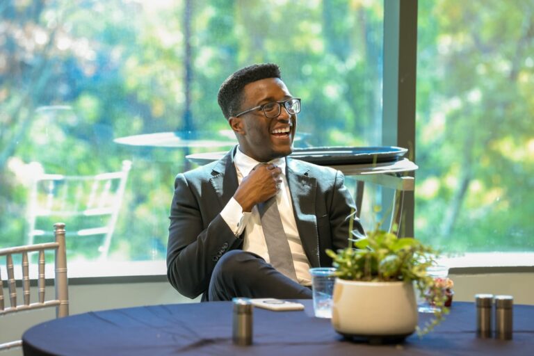 A man swearing glasses and a suit and tie smiles while seated at a table during the BECA Future Leaders Luncheon.