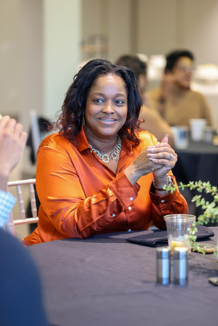 A woman in a bright orange shirt clasps her hands while listening to a colleague talk.