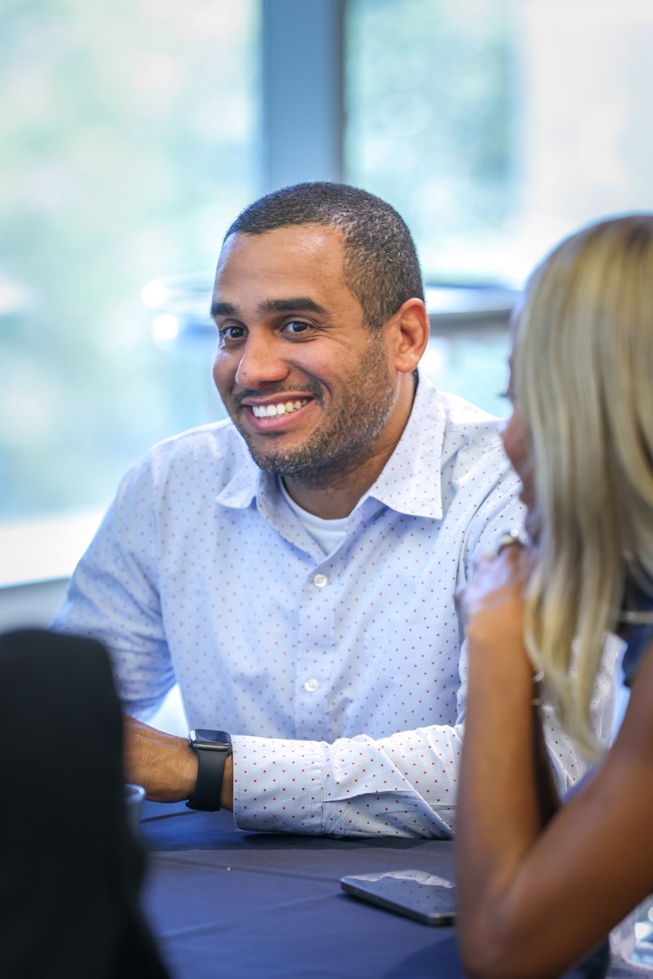 A man in a blue dress shirt with small dots on it smiles as he listens to a colleague at the BECA Future Leaders Luncheon.