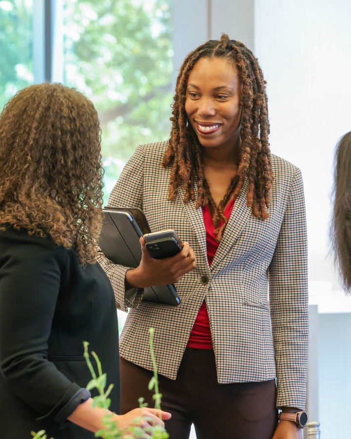 A woman wearing a checked blazer and a red top smiles and listens to a colleague.