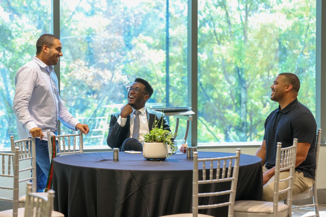 Three men engaged in conversation - one man is standing while two are seated. They are all smiling.