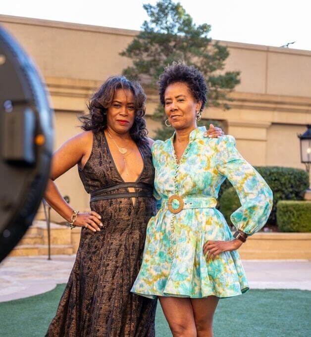 Two women in dresses pose for a photographer at an outdoor event.