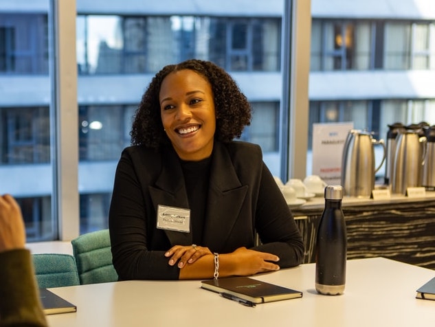 Smiling woman in a black blazer with a name tag, seated at a table in a modern office with a city view.
