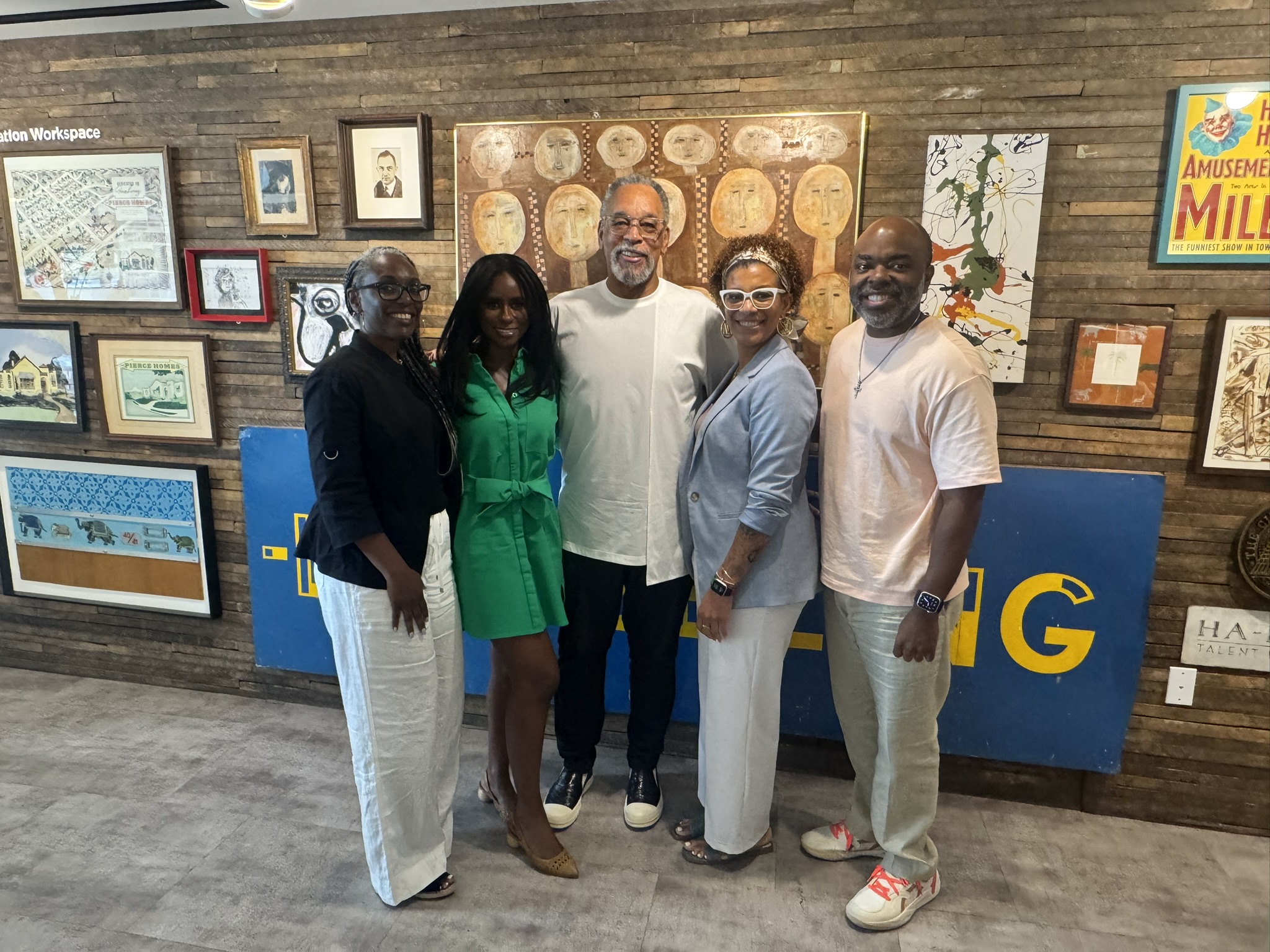 Five adults pose together in front of an eclectic gallery wall filled with colorful framed artwork and signage. They are smiling and dressed in stylish, business-casual attire, standing on a gray floor in what appears to be a creative or collaborative workspace.