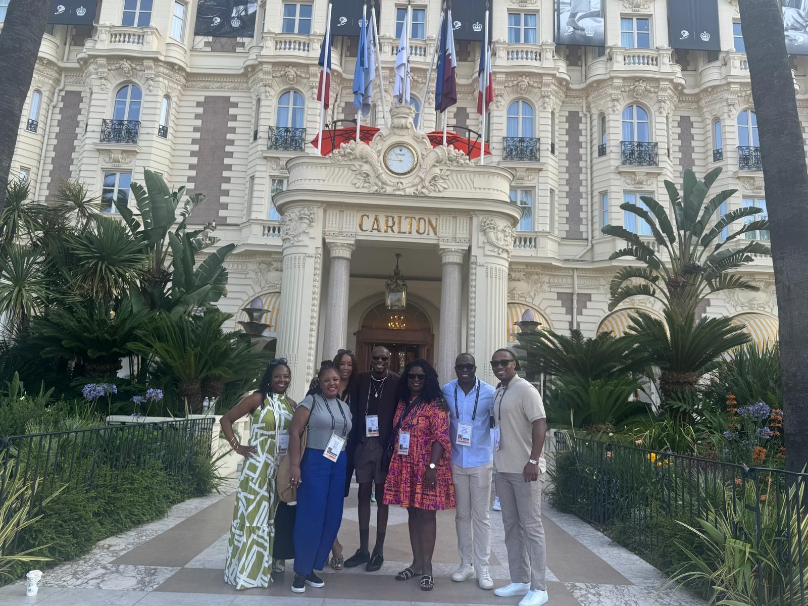 A group of seven people stand smiling in front of the grand entrance to the Carlton Hotel, framed by tall palm trees, lush greenery, and elegant architecture. The group wears conference badges, suggesting they’re attending a professional event, with the iconic hotel adding a touch of luxury to the moment.