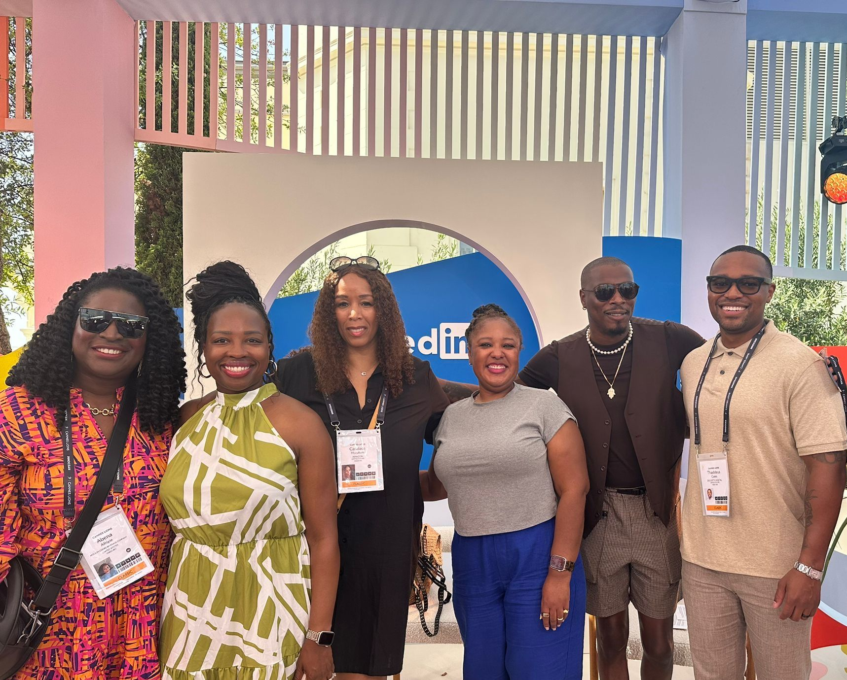 A group of seven pA group of seven professionally dressed people pose together and smile for a photo at a LinkedIn-branded event. The background features a modern, colorful setup with vertical slats and a lighted stage area, while several individuals wear event badges and hold microphones, suggesting participation in a panel or networking session.eople pose together at a LinkedIn-branded event space with a beach-themed backdrop featuring a sun and waves. Two women are seated in red-and-white striped deck chairs, while five others stand behind them. They are dressed in a mix of stylish and casual summer attire, with some wearing lanyards with event badges. A blue-and-white striped umbrella adds to the beach-like ambiance. The setting is outdoors with bright sunlight.