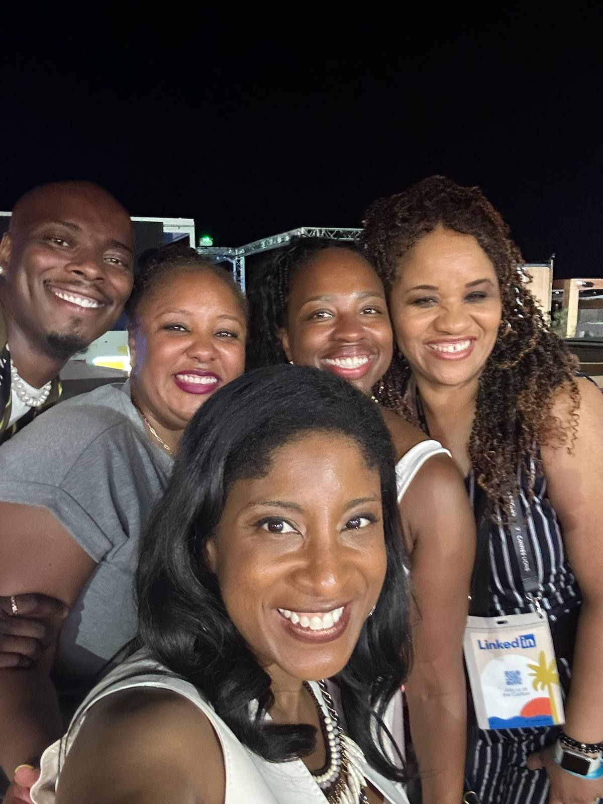 A group of six people smile brightly in a close-up selfie taken at night, with event lights and stage structures visible in the background. Everyone appears joyful and relaxed, with one attendee wearing a LinkedIn lanyard, indicating the photo was taken during after LinkedIn-hosted event or gathering.