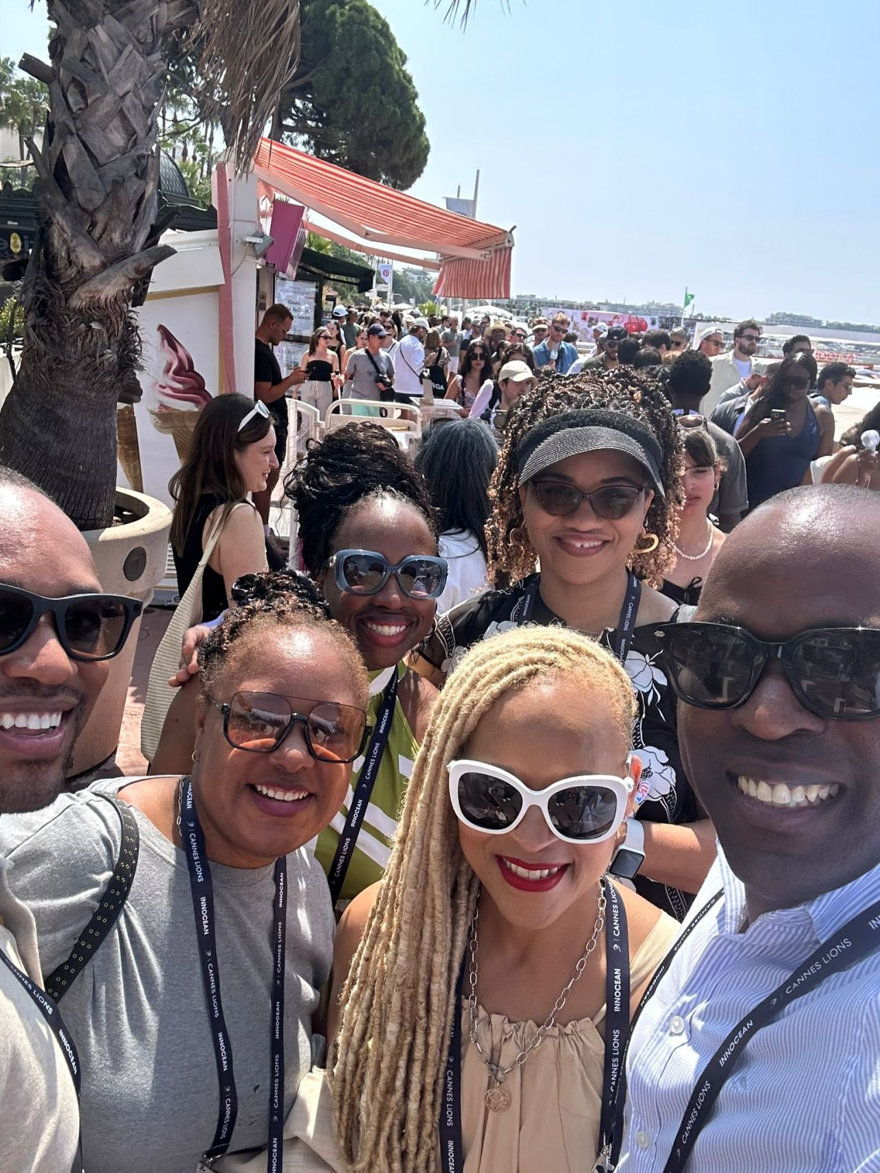 Six smiling attendees take a sunny selfie near a crowded boardwalk lined with palm trees, beachgoers, and a soft-serve ice cream stand. They wear lanyards marked “Cannes Lions,” suggesting they're part of the international festival crowd enjoying both professional networking and seaside leisure.