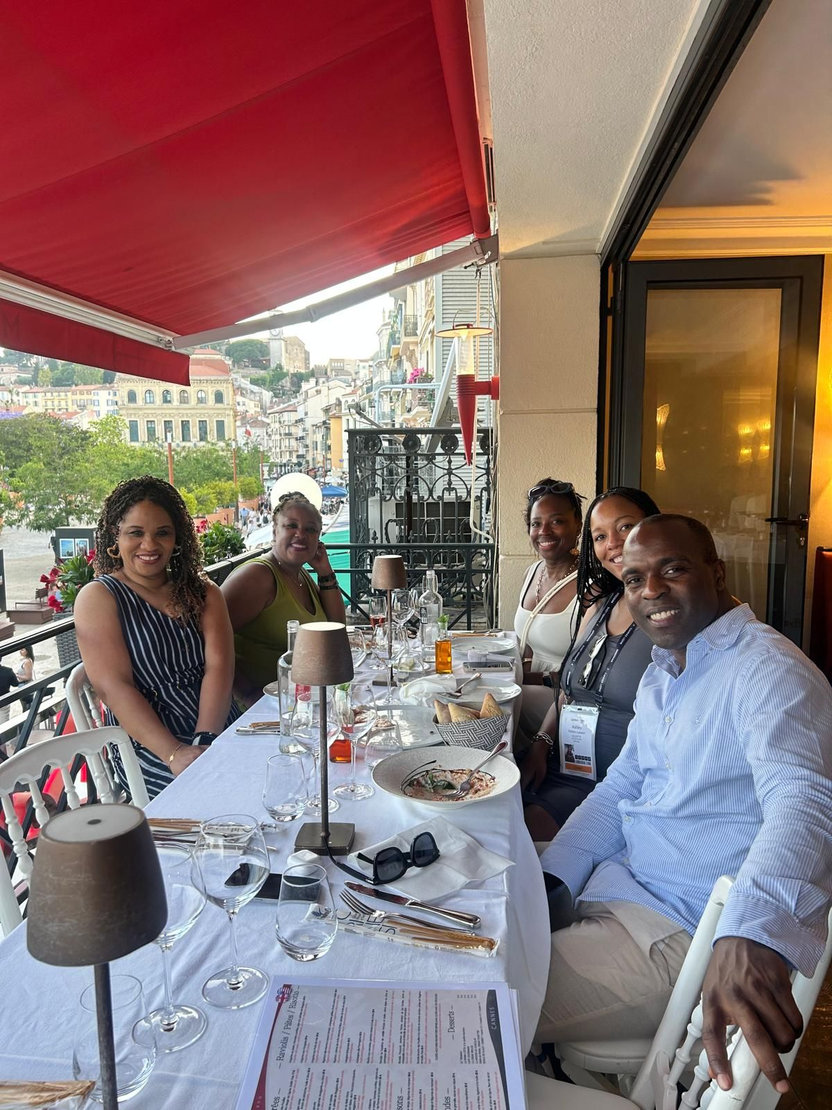 Six people smile and gather around a long white-tableclothed table at an outdoor restaurant balcony overlooking a charming European street. With wine glasses, menus, and a partially finished dish visible, the group appears to be enjoying a relaxed, upscale meal—likely as part of a professional event or celebration.